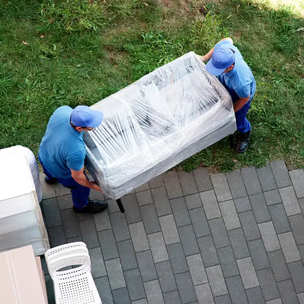 Zwei Möbelpacker tragen ein perfekt eingepacktes Sofa durch einen Garten zum Umzugswagen